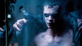 Bill Skarsgard is in The Crow, he has dark makeup around his eyes, tattooes on his chest, he is putting a dark trench coat on as he looks at himself in a mirror.