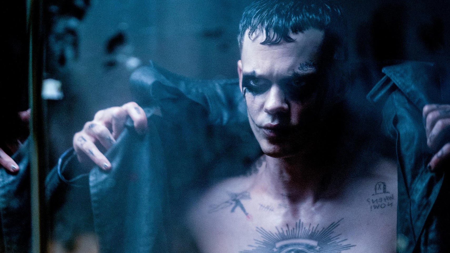Bill Skarsgard is in The Crow, he has dark makeup around his eyes, tattooes on his chest, he is putting a dark trench coat on as he looks at himself in a mirror. - 1