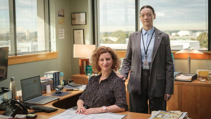 Two women in formal attire in an office, one is sat down, the other resting her hand on the back of a chair, in The Office Australia.
