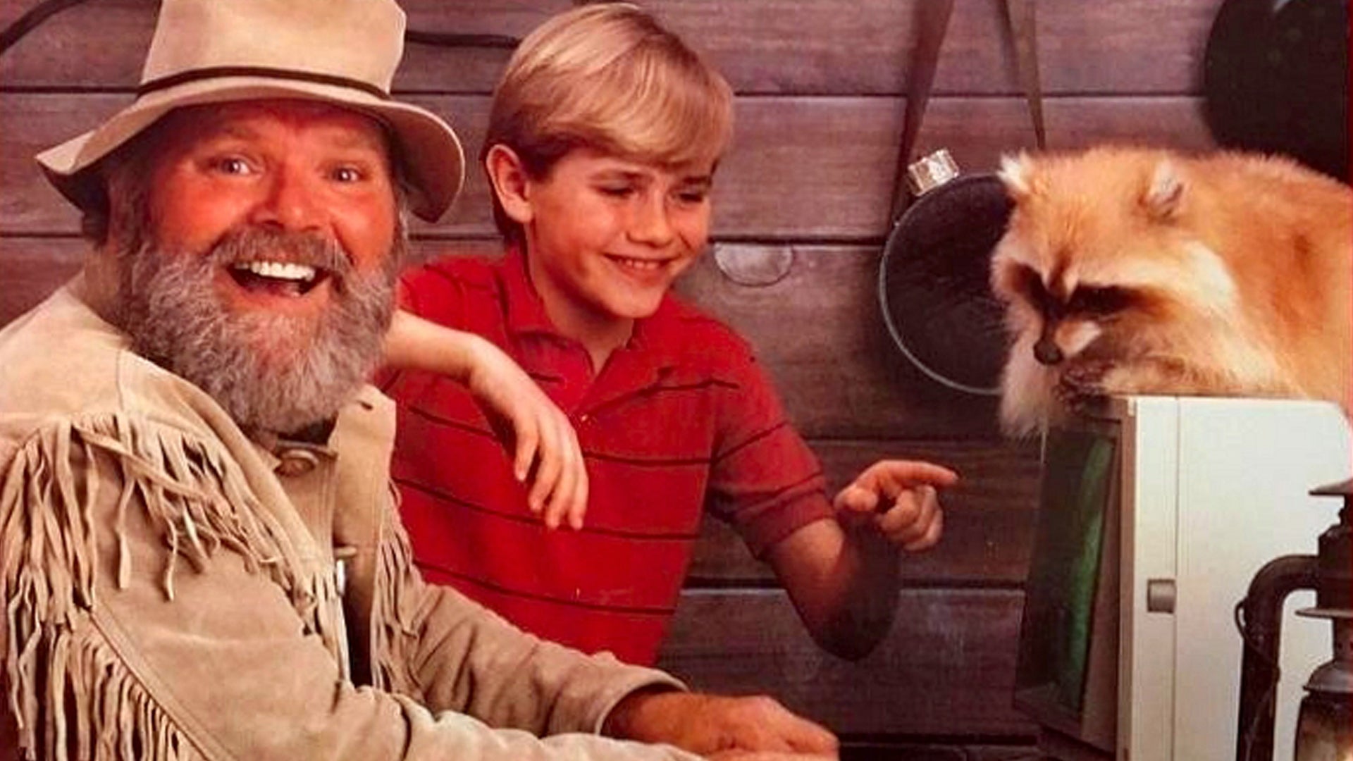 An old man wearing old timey clothes smiling sat at a computer with a boy pointing at the screen, a raccoon sat on top of it.