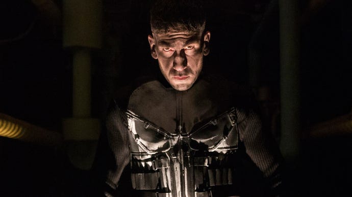 Jon Bernthal is stood as The Punisher in the show of the same name, he's looking into the camera, wearing a military-like uniform with a skull on it.