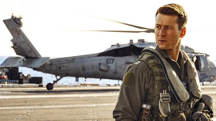 Glen Powell in Top Gun: Maverick, he’s stood in flight gear, a helicopter parked behind him.