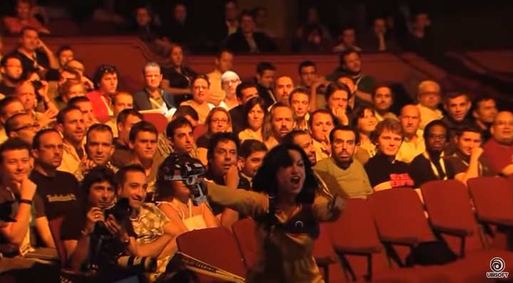 A woman in a yellow shirt with a laser tag gun and vest points and yells The theater audience sitting behind is full of people who appear either bored or bemused