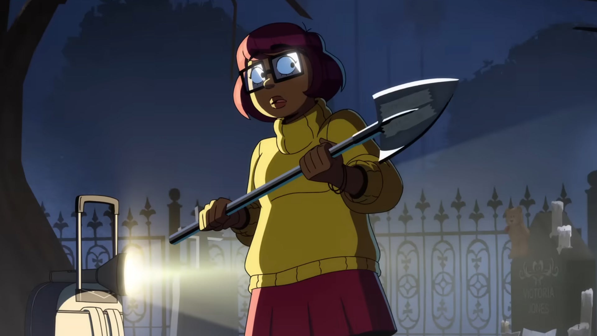 The titular Velma is holding a shovel in a graveyard, a flashlight lighting her up from the side, she looks slightly scared.