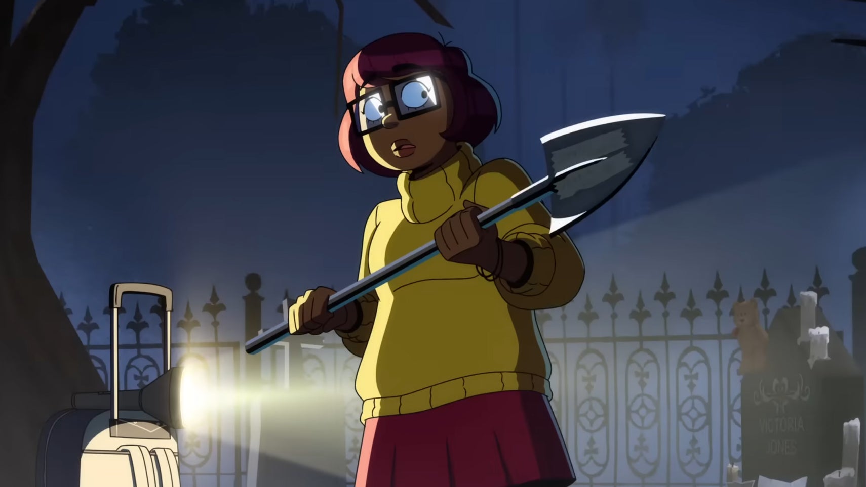 The titular Velma is holding a shovel in a graveyard, a flashlight lighting her up from the side, she looks slightly scared. - 1