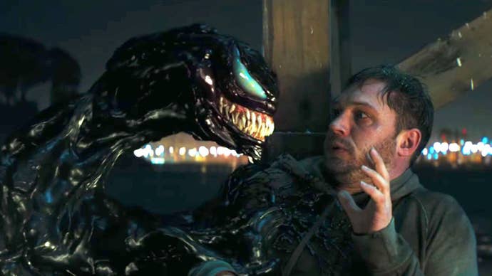 Tom Hardy as Eddie Brock is sat up against a metal beam, sweaty, holding his hand up in fear, as Venom looks at him smiling in 2018’s Venom.