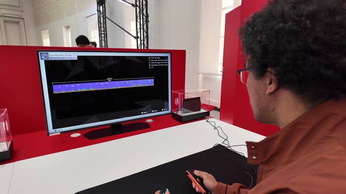 A man in a brown coat and dark hair plays the Nintendo Switch 2 on small monitor.