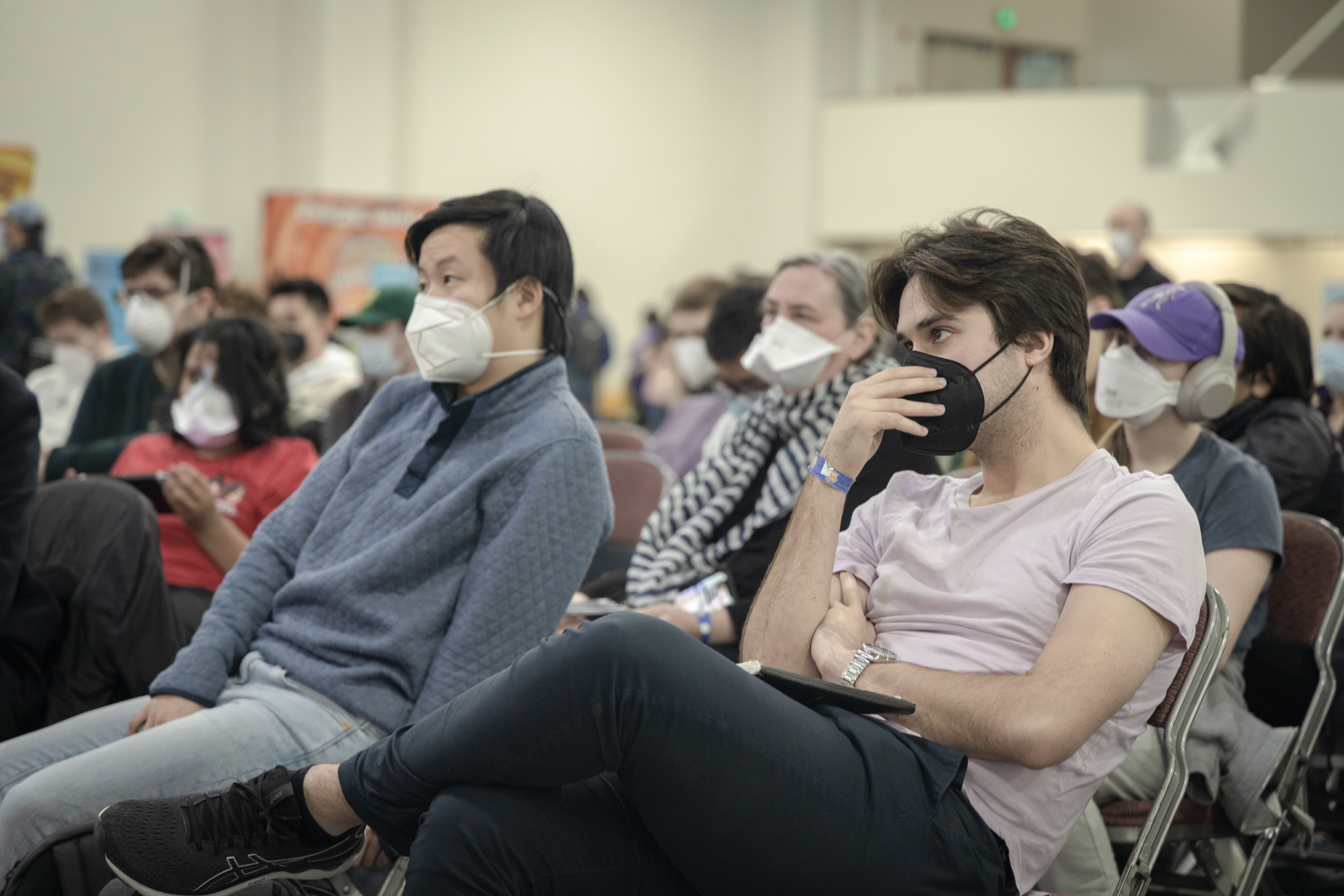 Pokemon champ Wolfe Glick lounging back in an auditorium chair, wearing a pinkish t-shirt and a black face mast. The audience around him is masked-up too. His friend and pundit Aaron Zheng sits next to him. They are staring intently at the stage (we can&rsquo;t see).