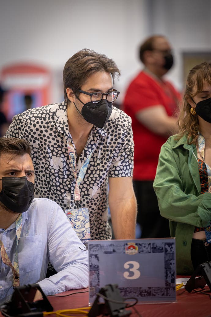 A photograph showing Pokemon champion Wolfe Glick spectating, and staring intently at action the camera is now showing. A black face mask and thick framed glasses cover a lot of his face.