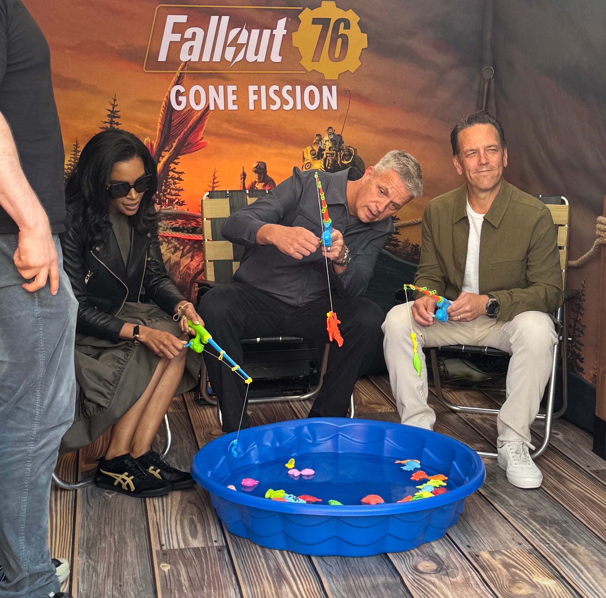 From left to right Sarah Bond, Matt Booty, and Phil Spencer sit around a small blue plastic paddling pool with toy fish in it holding toy rods, at the Fallout 76 Gone Fission SGF 2025 booth.