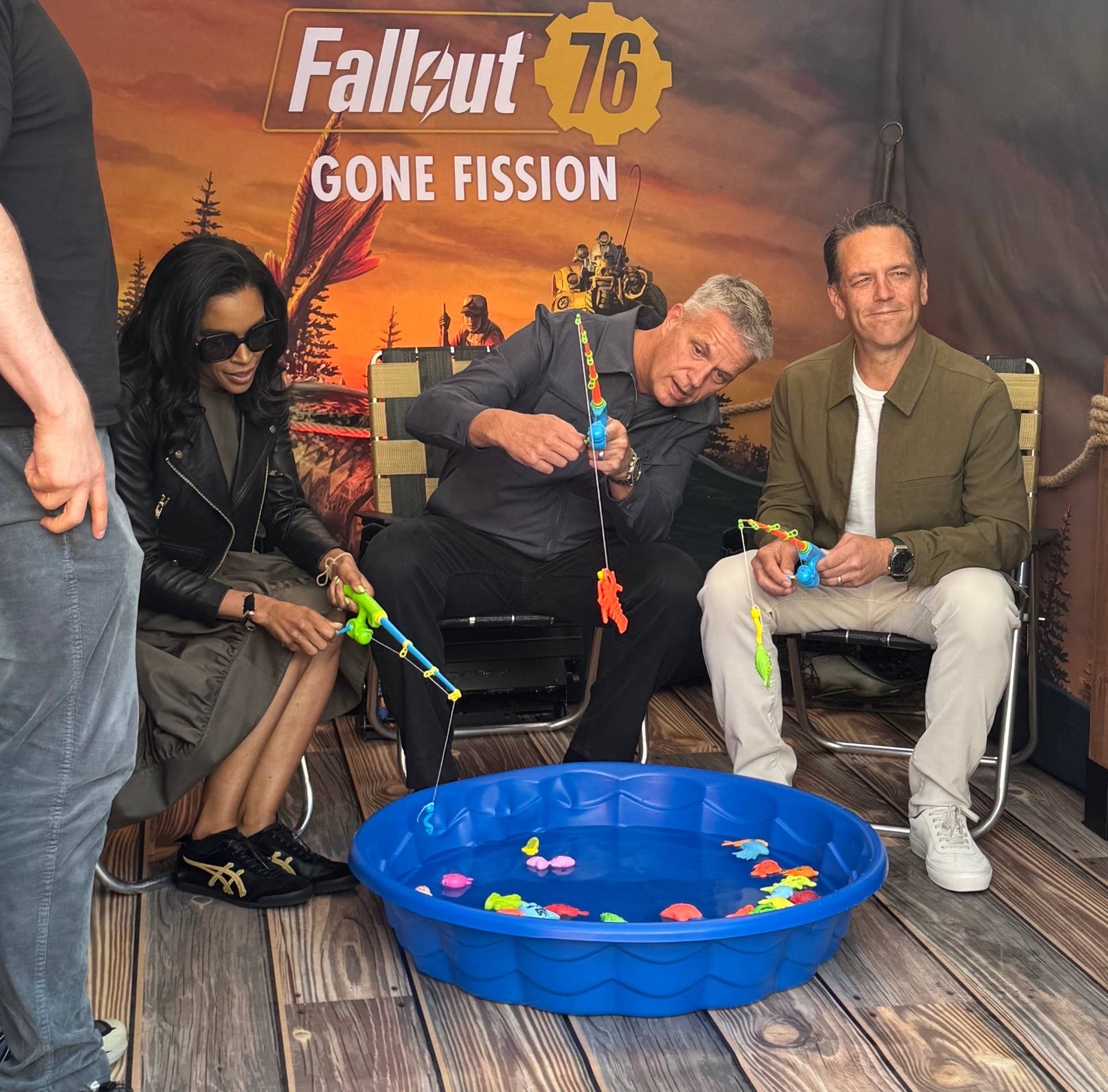 From left to right Sarah Bond, Matt Booty, and Phil Spencer sit around a small blue plastic paddling pool with toy fish in it holding toy rods, at the Fallout 76 Gone Fission SGF 2025 booth.
