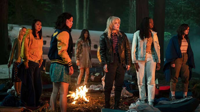 Various Yellowjackets cast members stood around a campfire looking at something off to the side.