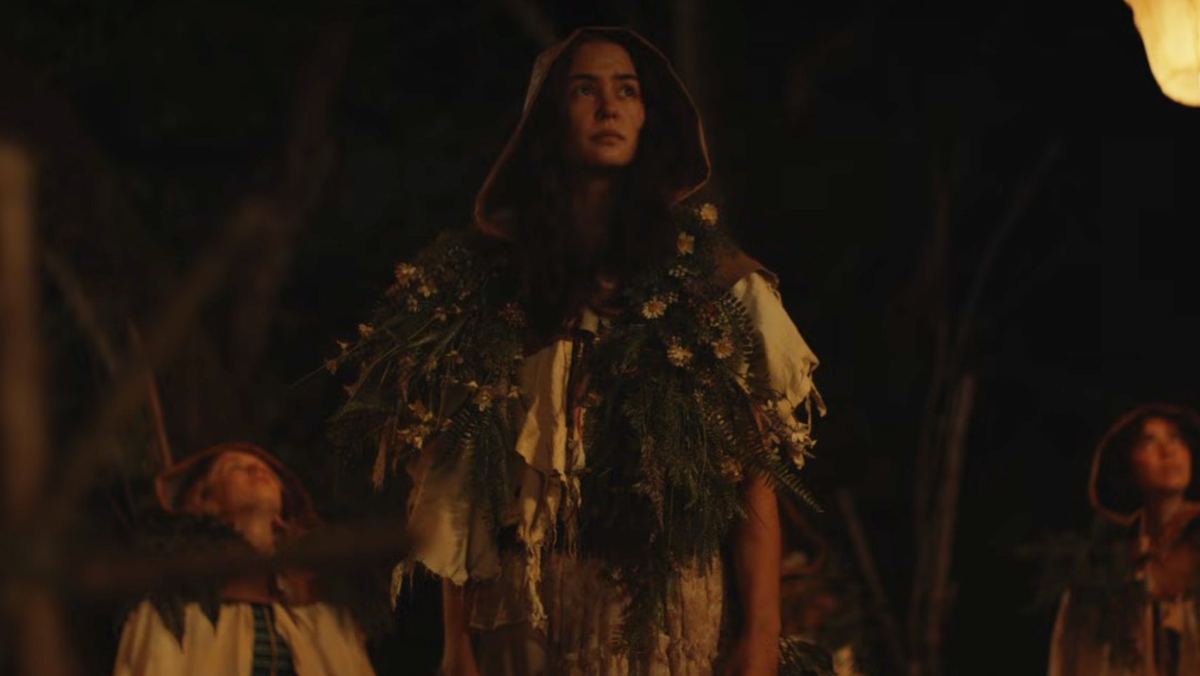 A young woman is wearing makeshift clothing in the woods at nighttime, lanterns flying into the sky, other young women nearby in Yellowjackets season 3. - 1