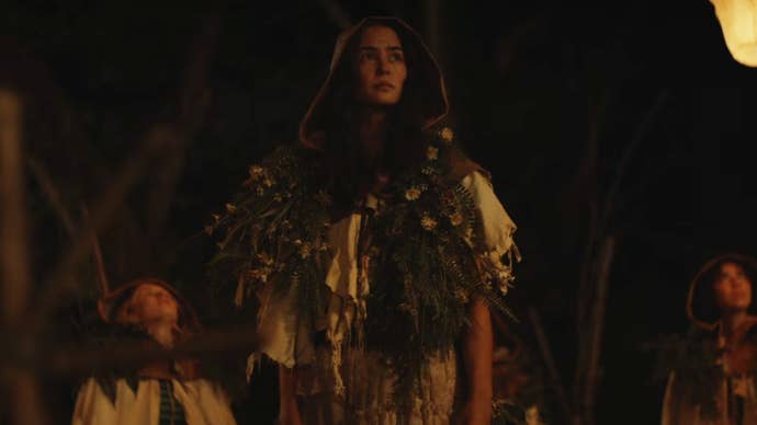 A young woman is wearing makeshift clothing in the woods at nighttime, lanterns flying into the sky, other young women nearby in Yellowjackets season 3.